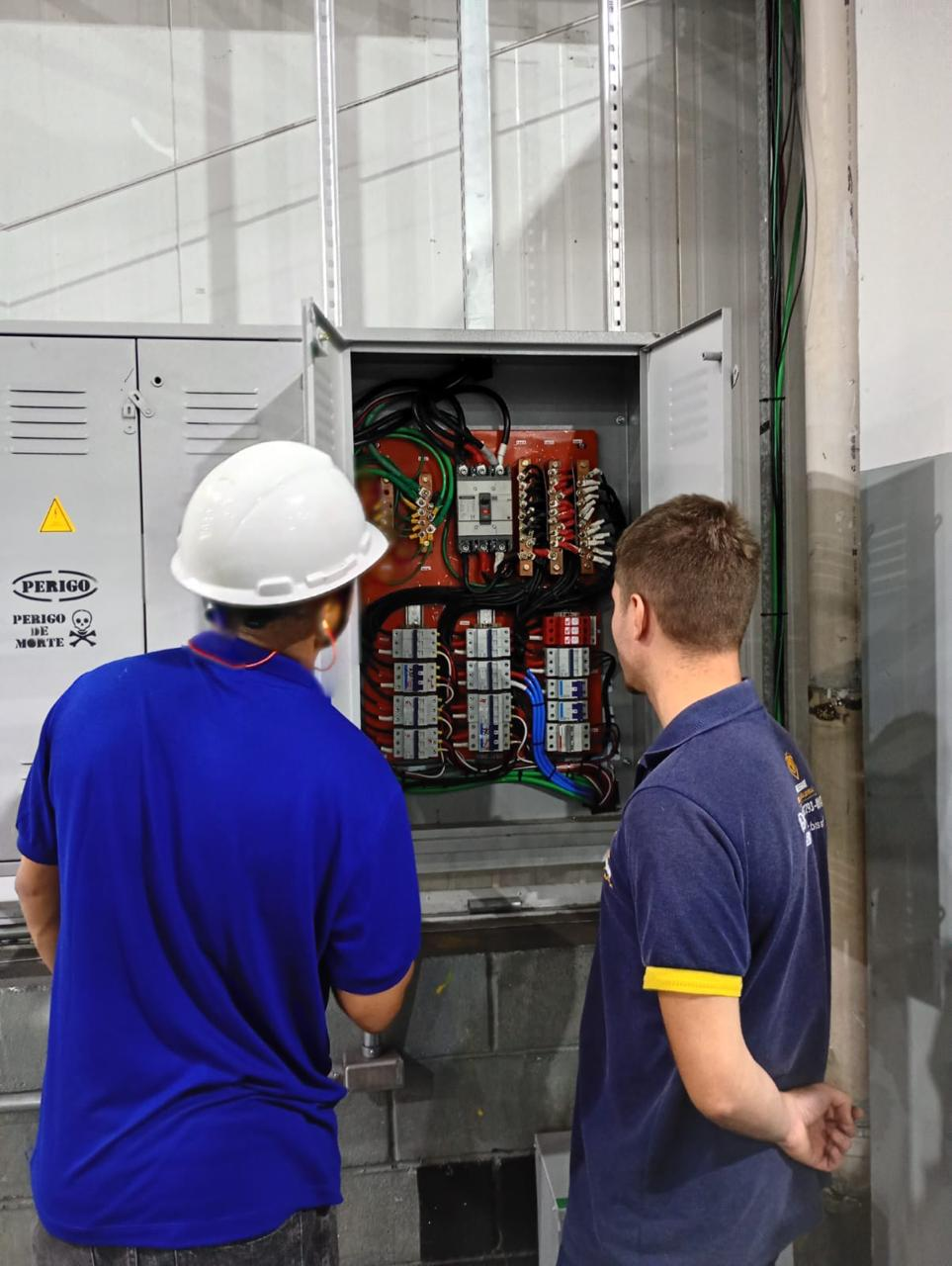 Dois eletricistas inspecionando quadro elétrico industrial trifásico com contatores e fiação organizada